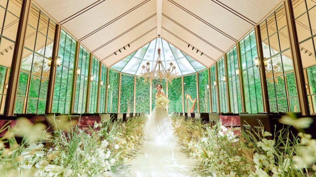 Bride in a white wedding gown holding a bouquet stands at the end of a glass-walled aisle lined with flowers and greenery, chandeliers overhead.