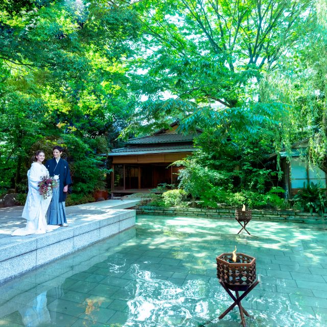 Bride in a white kimono and groom in dark formal wear stand on stone steps beside a shallow pool, surrounded by lush trees.