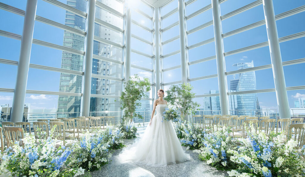 Bride in a white wedding dress holding a bouquet stands in a glass atrium, surrounded by blue and white flowers with a city skyline outside the windows.
