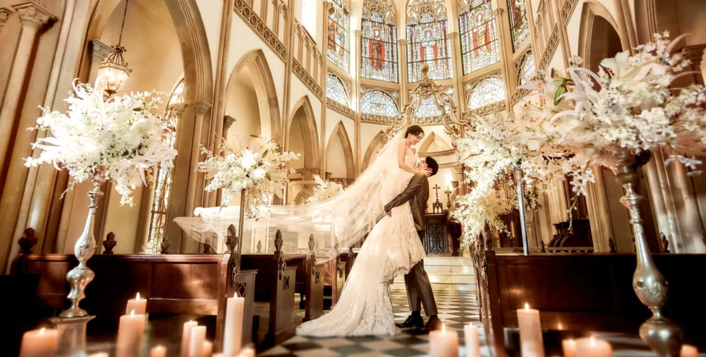 Bride and groom embrace in a grand church, veil flowing as she is lifted, with stained-glass windows behind.
