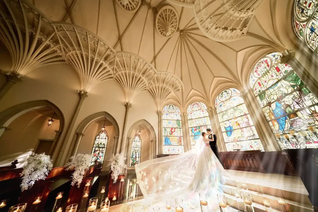 Bride and groom kiss in a grand church, with a long veil sweeping the aisle and colorful stained-glass windows behind them.