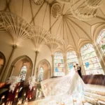 Bride and groom kiss in a grand church, with a long veil sweeping the aisle and colorful stained-glass windows behind them.