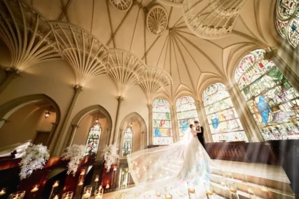 Bride and groom kiss in a grand church, with a long veil sweeping the aisle and colorful stained-glass windows behind them.