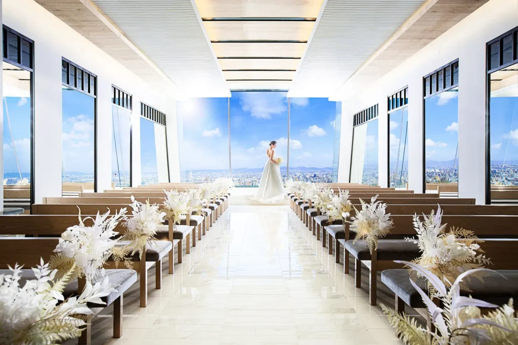 Bride in a white wedding gown stands at the end of a bright chapel, with white floral arrangements along the pews and a city skyline outside.